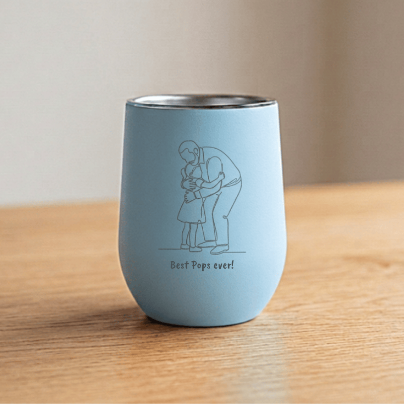 A light blue insulated stemless wine tumbler with a stainless steel rim, featuring a simple line drawing of a father and child hugging with the text 'Best Pops ever!' printed below, set on a wooden table.